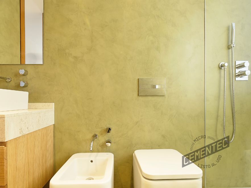 Image showing a green microcement wall, a toilet, a bidet, a wooden washbasin and a shower in what is an example of a microcement and wood bathroom. 